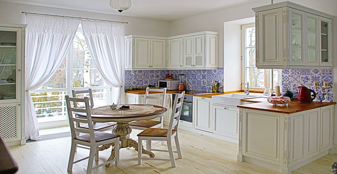 Biała kuchnia drewniana na wymiar. Meble kuchenne w tym kolorze ładnie współgrają z brązowymi blatami i z niebieskimi kaflami na ścianach.