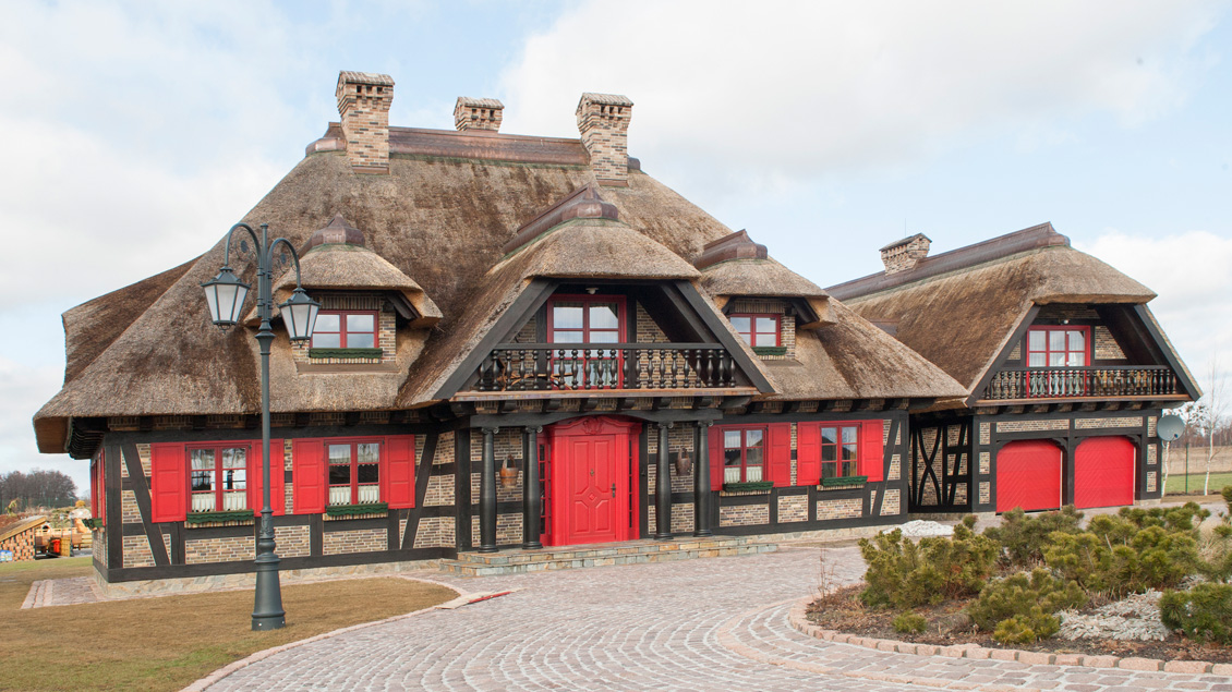 Budynek zajazdu z drewnianymi oknami wykonanymi na wymiar - okna z drewnianymi czerwonymi okiennicami, na zdjęciu też czerwone drewniane drzwi zewnętrzne, mur pruski, dom kryty strzechą.