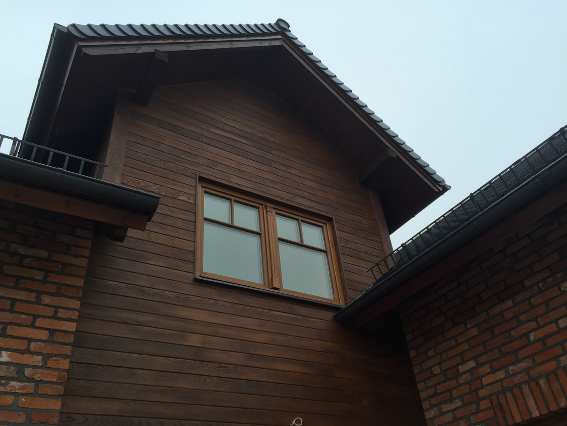 Drewniane okno na wymiar wkomponowane w brązową obłożoną drewnem ścianę domku jednorodzinnego.