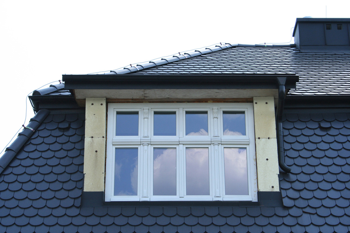 Drewniane białe okno zdobione pilastrami / półkolumnami na poddaszu domu, widok okna z zewnątrz na tle czarnego dachu.